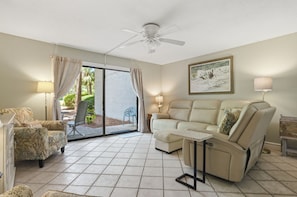A living room with a sofa, armchair, and a table, a ceiling fan, and a door leading to a balcony.