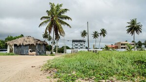 A rural setting with a thatched-roof hut, palm trees, and a modern building in the background. - Apto c Piscina e rea Gourmet a 100m da Praia (Japaratinga)
