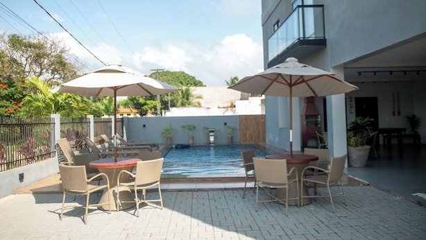 An outdoor pool area with wicker furniture and umbrellas, surrounded by a brick patio and a building with a balcony. - Apto c Piscina e rea Gourmet a 100m da Praia (Japaratinga)