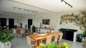 A spacious dining area with a wooden table and wicker chairs, surrounded by potted plants. - Apto c Piscina e rea Gourmet a 100m da Praia (Japaratinga)