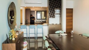 Un coin repas avec une longue table et des chaises bleues, un espace cuisine où une personne est en train de préparer un plat, et un verre à vin posé sur la table.