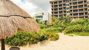 Une plage avec un parasol en chaume, une végétation luxuriante et, au loin, un immeuble d’hôtel à plusieurs étages.