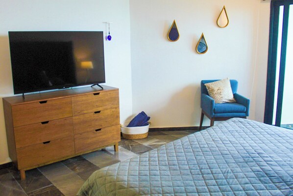 Une chambre à coucher comprenant un lit, une commode en bois, un téléviseur, une chaise bleue et un mur orné de deux objets décoratifs.