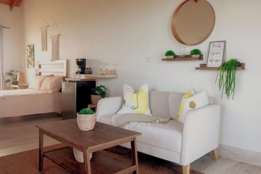 Ein gemütliches Wohnzimmer mit einem weißen Sofa, einem runden Spiegel und einem Couchtisch. Regale mit Pflanzen und ein Schild mit der Aufschrift 'Truth Love Hope'.