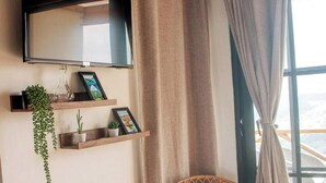 A room with a mounted TV, wooden shelves with plants and framed pictures, and a window with curtains. - Cabin in the mountains w/ Breakfast - Cabin 1 (Miches)