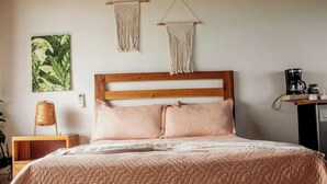 A bedroom with a wooden headboard, a bed with a pink quilt, two macramé wall hangings, a green leafy painting, a wicker chair, and a coffee machine on a shelf. - Cabin in the mountains w/ Breakfast - Cabin 1 (Miches)