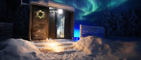 Un edificio imbiancato dalla neve con un'ghirlanda sulla porta, un cartello con il numero 24, e vista sull'aurora boreale.