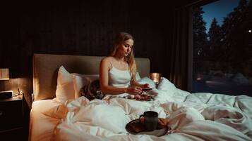 Una persona sentada en una cama con un plato de comida y una taza, en una habitación con ventana que da a árboles.