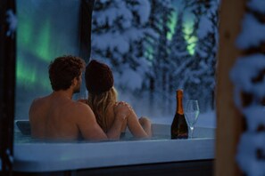 Two people in a hot tub with a bottle of champagne and a glass, surrounded by a snowy landscape.