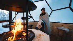 Una persona envuelta en una manta, sentada junto a una fogata dentro de una estructura en forma de domo geodésico, con vista a árboles cubiertos de nieve.
