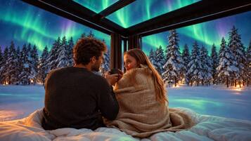 Una pareja sentada en una cama con vistas a las auroras boreales a través de una ventana, rodeada de árboles cubiertos de nieve.