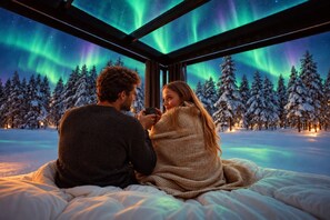 Una coppia seduta su un letto con vista sull'aurora boreale attraverso una finestra, circondata da alberi coperti di neve.