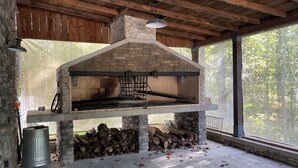 An outdoor stone fireplace with a chimney, surrounded by logs and a metal trash can.