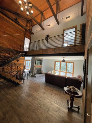 A spacious loft with a wooden staircase, a fireplace, a large window, and a high ceiling.