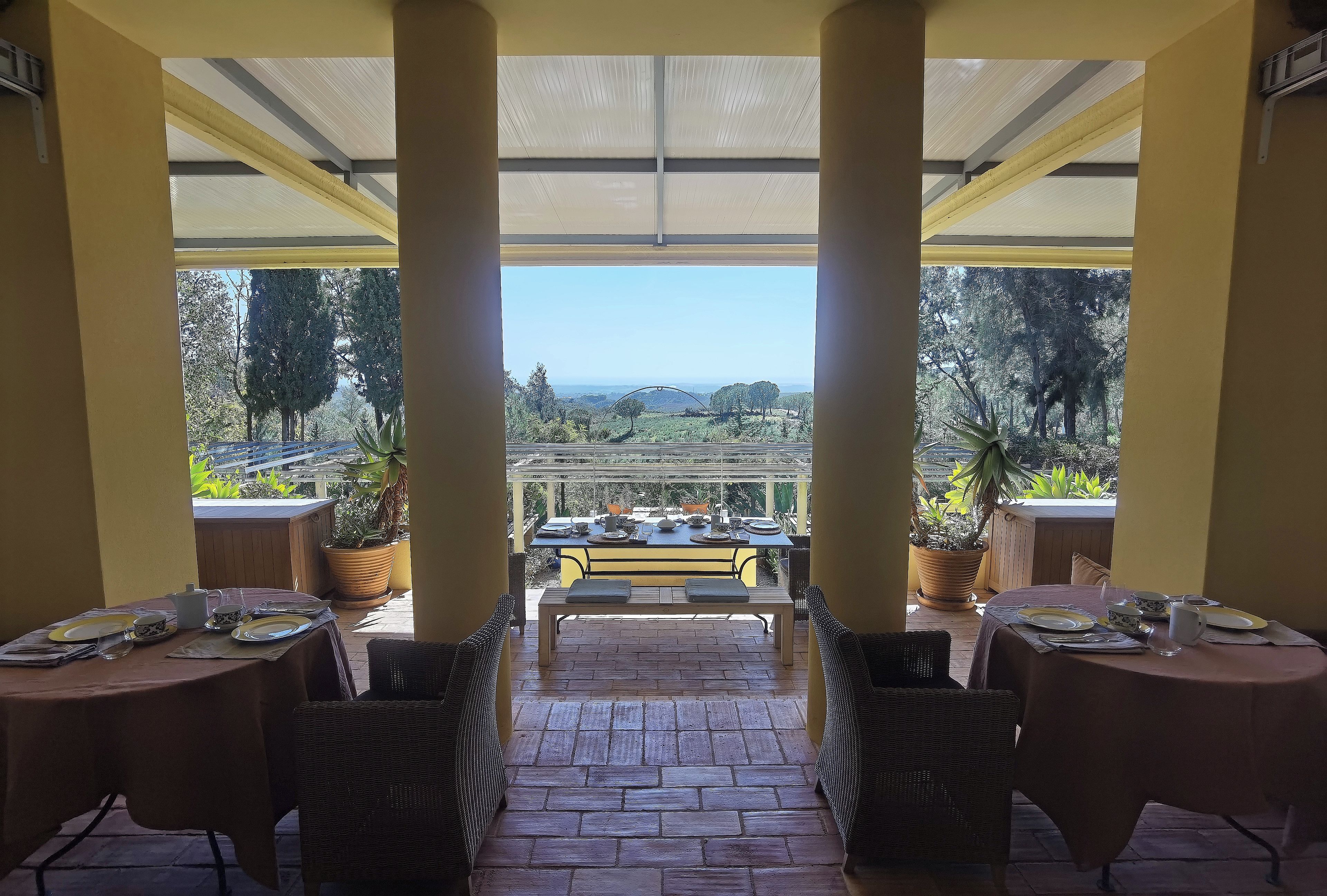 Un espacio exterior cubierto para comer con mesas dispuestas para dos personas, vistas del paisaje y plantas en macetas.