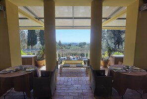 Un espace extérieur abrité, aménagé pour les repas, avec des tables pour deux personnes, une vue sur le paysage et des plantes en pot.