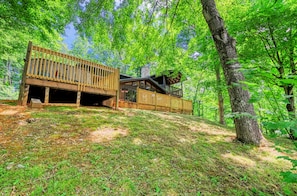 A cabin with a wooden deck, a covered porch, and a stone foundation, surrounded by a forested hillside.