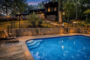 A swimming pool with a stone wall and a wooden deck, surrounded by a wooden fence and trees.