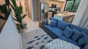 A modern living room with a blue sofa, a kitchen area with a refrigerator and microwave, and a dining table with chairs. - Stylish 2 bed condo with mountain & pool view (Jaco)