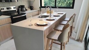 A modern kitchen with a central island, bar stools, and a window providing a view of greenery. - Stylish 2 bed condo with mountain & pool view (Jaco)