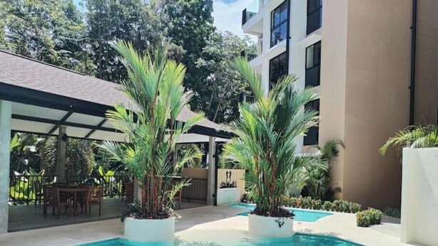 A swimming pool with surrounding greenery and a covered seating area. - Stylish 2 bed condo with mountain & pool view (Jaco)