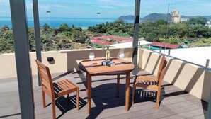A covered outdoor seating area with a table and chairs, overlooking a coastal landscape with buildings and greenery. - Stylish 2 bed condo with mountain & pool view (Jaco)