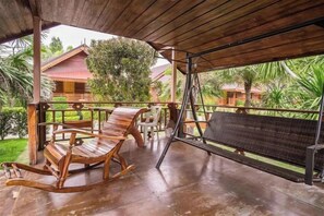 A covered outdoor area with wooden rocking chairs and a bench, a wooden table, and a view of a red-roofed building in the background. - Ruentara Resort & Villa (Buri Ram)