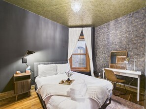 A bedroom with a bed, bedside table, lamp, chair, desk, and window.
