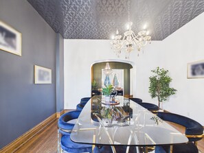 A dining area with a glass table, blue upholstered chairs, a chandelier, and a view into another room.