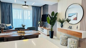 Una sala de estar moderna con comedor, que cuenta con un gran ventanal con cortinas translúcidas, un televisor de pantalla plana, un espejo redondo y una planta en maceta.