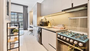 A modern kitchen with a gas stove, a built-in oven, and a dishwasher. There is a window with curtains, a small seating area, and a plant on a shelf. - Posh Studio in MBR by Deleguest (Dubai)