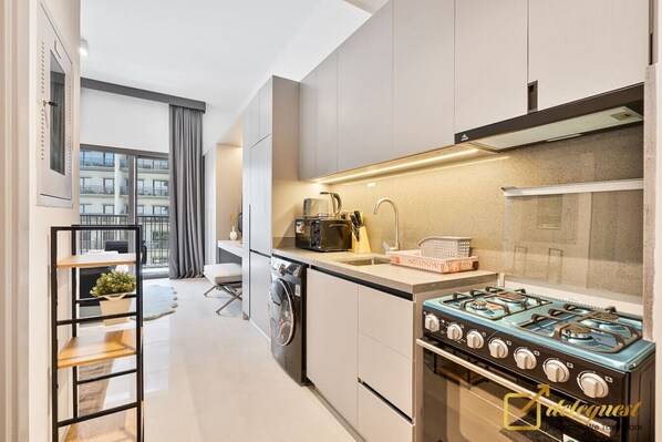 A modern kitchen with a gas stove, a built-in oven, and a dishwasher. There is a window with curtains, a small seating area, and a plant on a shelf.