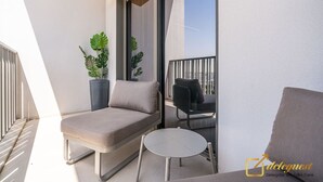 A balcony with a sofa, a small table, and two chairs. There are potted plants and a view of the city. - Posh Studio in MBR by Deleguest (Dubai)