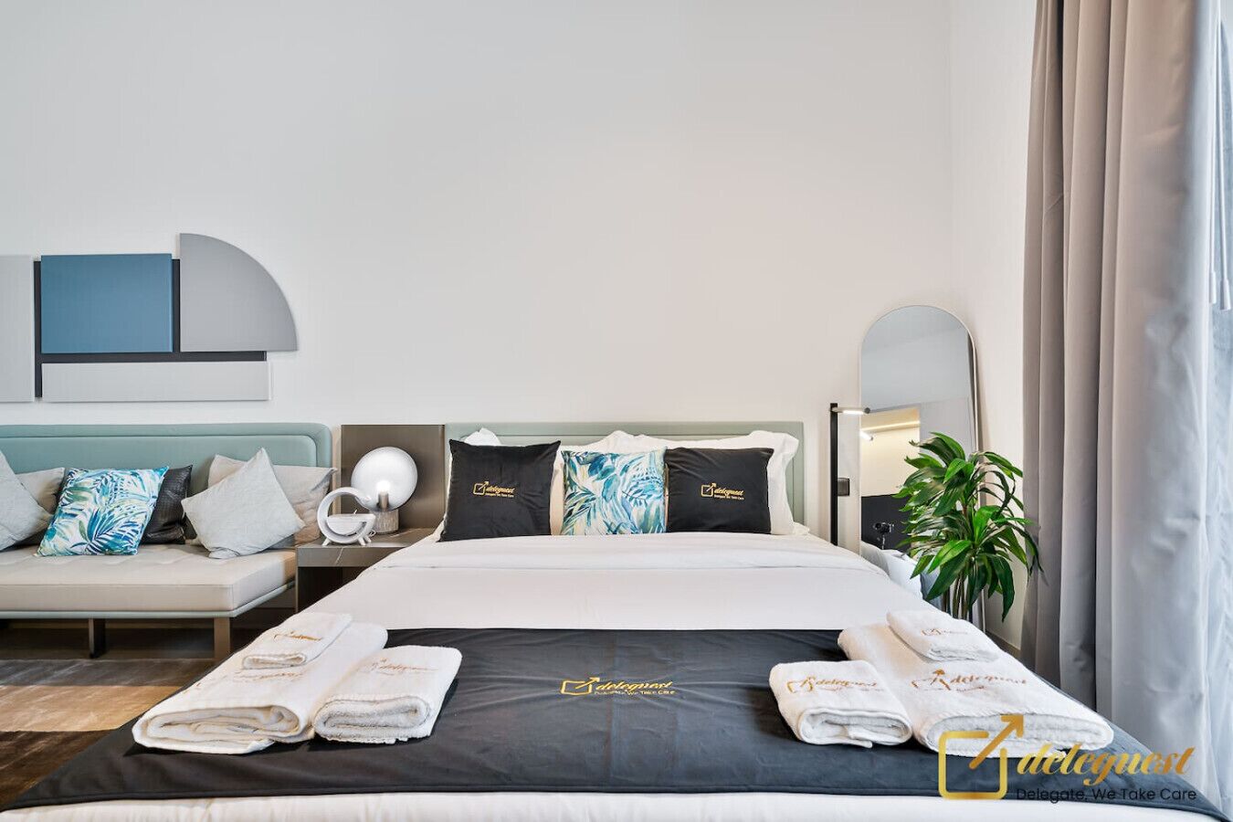 Ein Hotelzimmer mit Bett, Sofa, Nachttisch, Lampe, Spiegel und einer Zimmerpflanze.