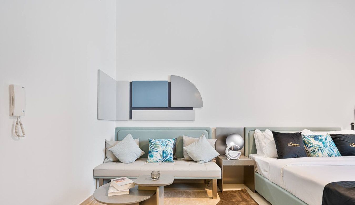 A hotel room with a sofa, a bed, a small table, and a nightstand. There is a phone on the wall and a logo on the bed.