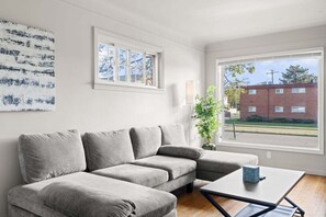 Olohuoneessa on harmaa kulmasohva, pieni pöytä, lamppu ja suuri ikkuna, josta avautuu näkymä tiilitaloon.