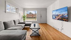 Una sala de estar moderna con un sofá seccional gris, una mesa de centro y un televisor grande montado en la pared que muestra un paisaje urbano.