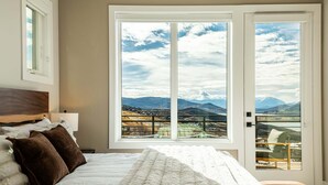 Un dormitorio con un ventanal grande, una cama con ropa de cama blanca y vistas a montañas y un lago.