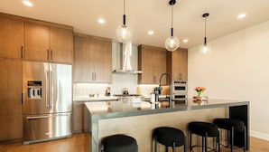 Una cocina moderna con electrodomésticos de acero inoxidable, armarios de madera, una isla central con taburetes altos y luces colgantes.