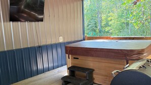 A wooden cabin with a hot tub, a television mounted on the wall, and a view of the forest. - Cozy Cabin with hot tub, just minutes from Nolin lake and Mammoth Cave park! (Leitchfield)