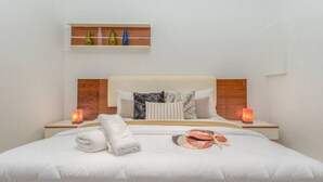 A neatly made bed with white linens, a folded towel, and a decorative item on the bed. There are two bedside tables with candles. Above the bed, there is a shelf with decorative vases. - Sirene 1-BR Apt Pool & Gym in Gemmayze (Beirut)