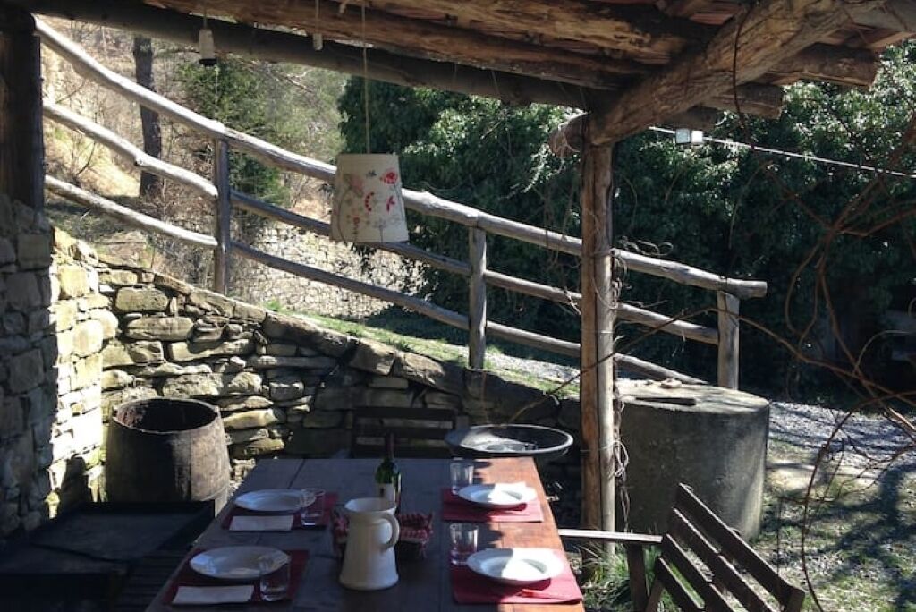 Un espace de restauration en plein air avec des murs en pierre, une table en bois dressée pour deux personnes et une pergola en bois rustique au-dessus.