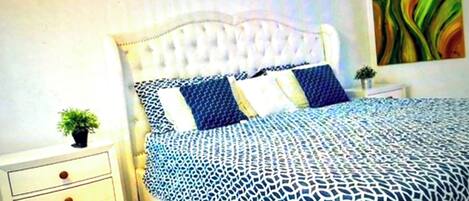Een slaapkamer met een gestoffeerd hoofdeinde, een bed met een decoratieve spreidek en een nachtkastje met lades.