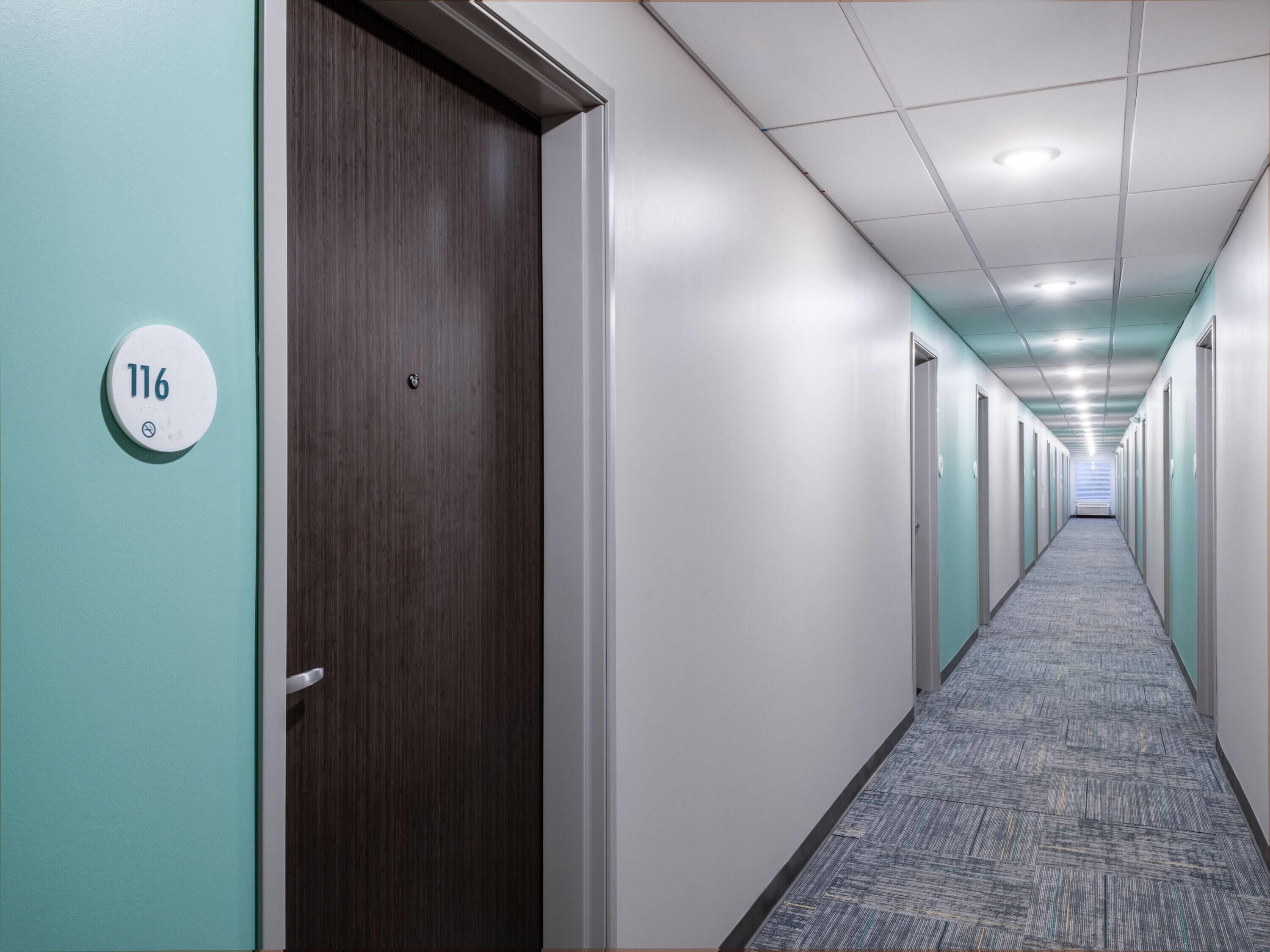 A hallway with teal walls, a carpeted floor, and doors numbered 116.