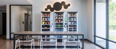 Un comedor moderno con una mesa larga y cuatro sillas, una máquina expendedora de aperitivos y un ventanal que ofrece vistas al exterior.
