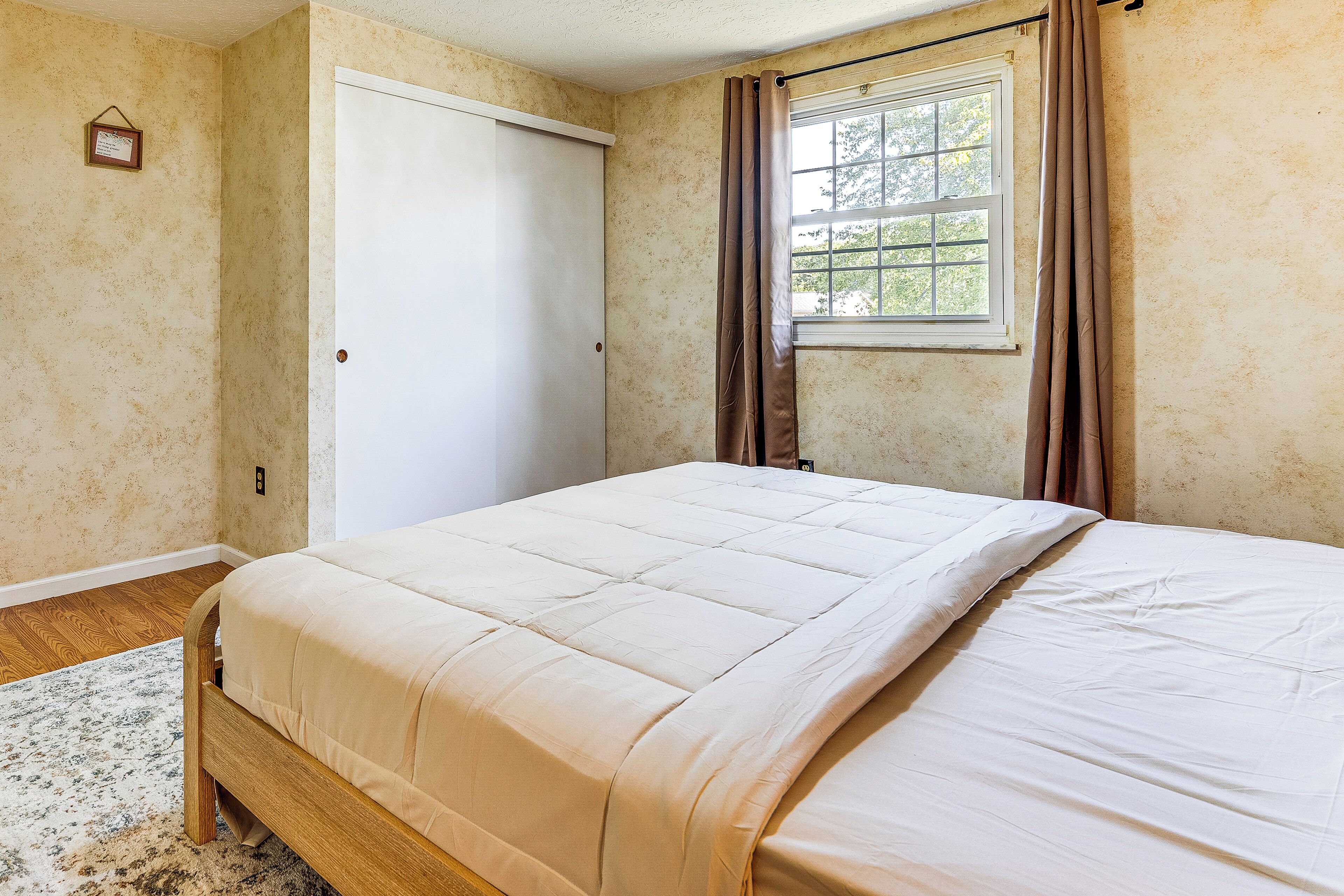 Un dormitorio con cama, ventana con cortinas, una puerta y un pequeño cuadro en la pared.