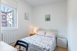 A bedroom with a bed, a chair, a nightstand with a lamp, a window, and artwork on the walls.