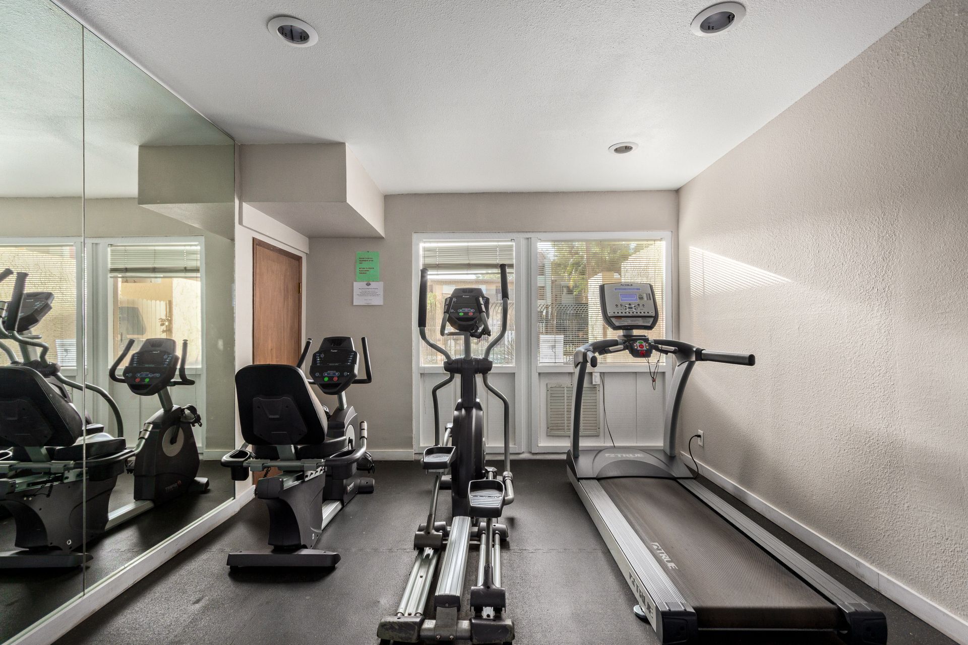 Un gimnasio con diverso equipo de ejercicio, incluyendo cintas de correr y bicicletas estáticas, un muro con espejos y una puerta que da a otra sala.