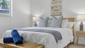 Un lit bien fait, avec du linge de lit blanc, un plaid bleu et des coussins à motifs. On aperçoit un banc en bois avec une serviette bleue à côté. Deux tables de chevet avec des lampes sont visibles.
