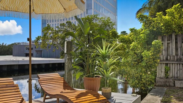 Une terrasse en bois, dotée de chaises longues et d’un parasol à motifs, entourée d’une végétation luxuriante et bordée d’un canal.